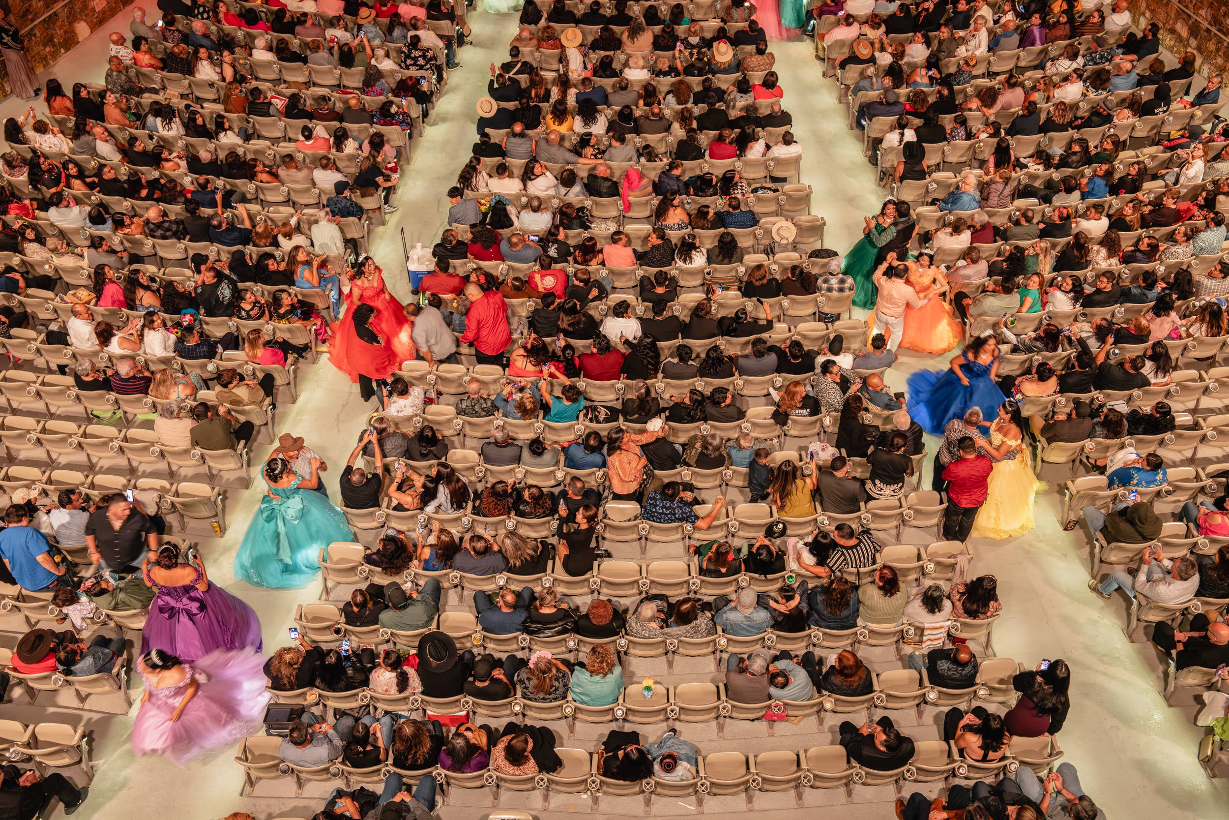 Crowd packed in theater seats at a colorful performance with dancers in blue, green, and pink skirts along the aisles
