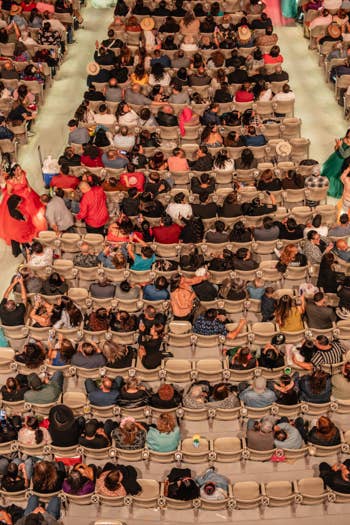 Crowd packed in theater seats at a colorful performance with dancers in blue, green, and pink skirts along the aisles