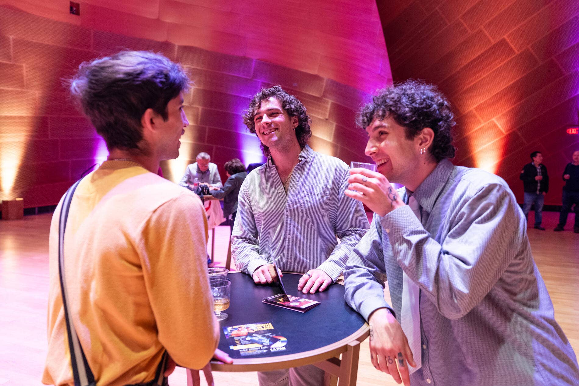 People mingle at a banquet bar, pouring drinks at a table with sparkling bottles under colorful stage lights.