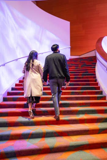 Two people walk hand in hand up red patterned theater stairs, lit by colorful stage projections.