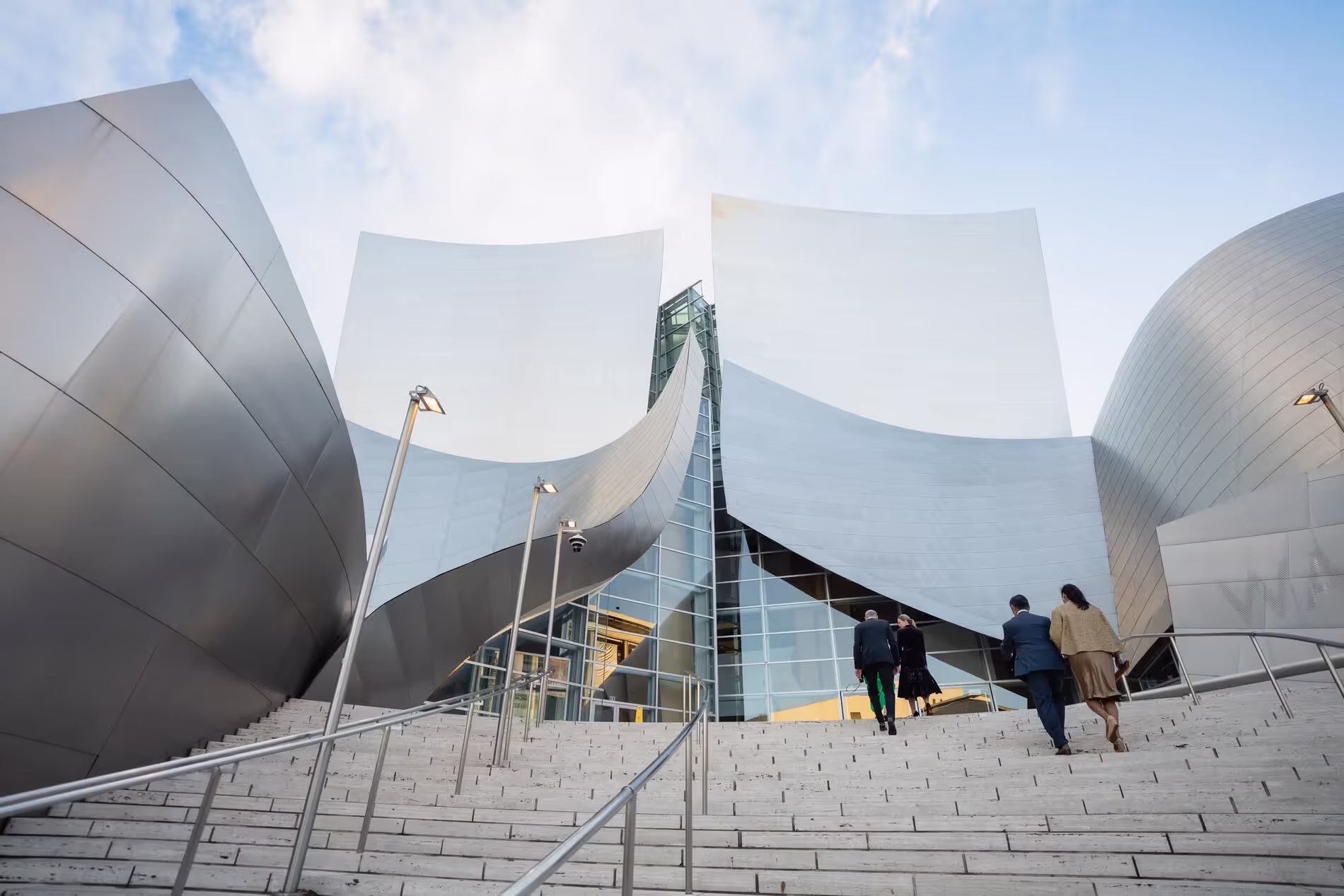 Modern walt disney concert hall building with curved white metal panels, steps, streetlights, and people walking at dusk