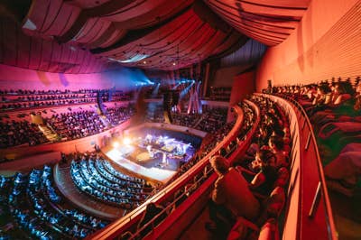 Audience fills a concert hall as musicians perform on stage under colorful red and blue lights