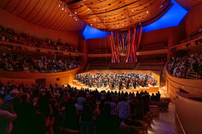 Concert hall interior with large orchestra on stage, audience seated, and organ pipes lit in blue and red.