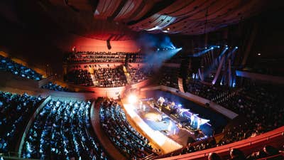 Concert hall with a stage illuminated by bright lights as performers and a large audience fill tiered seating