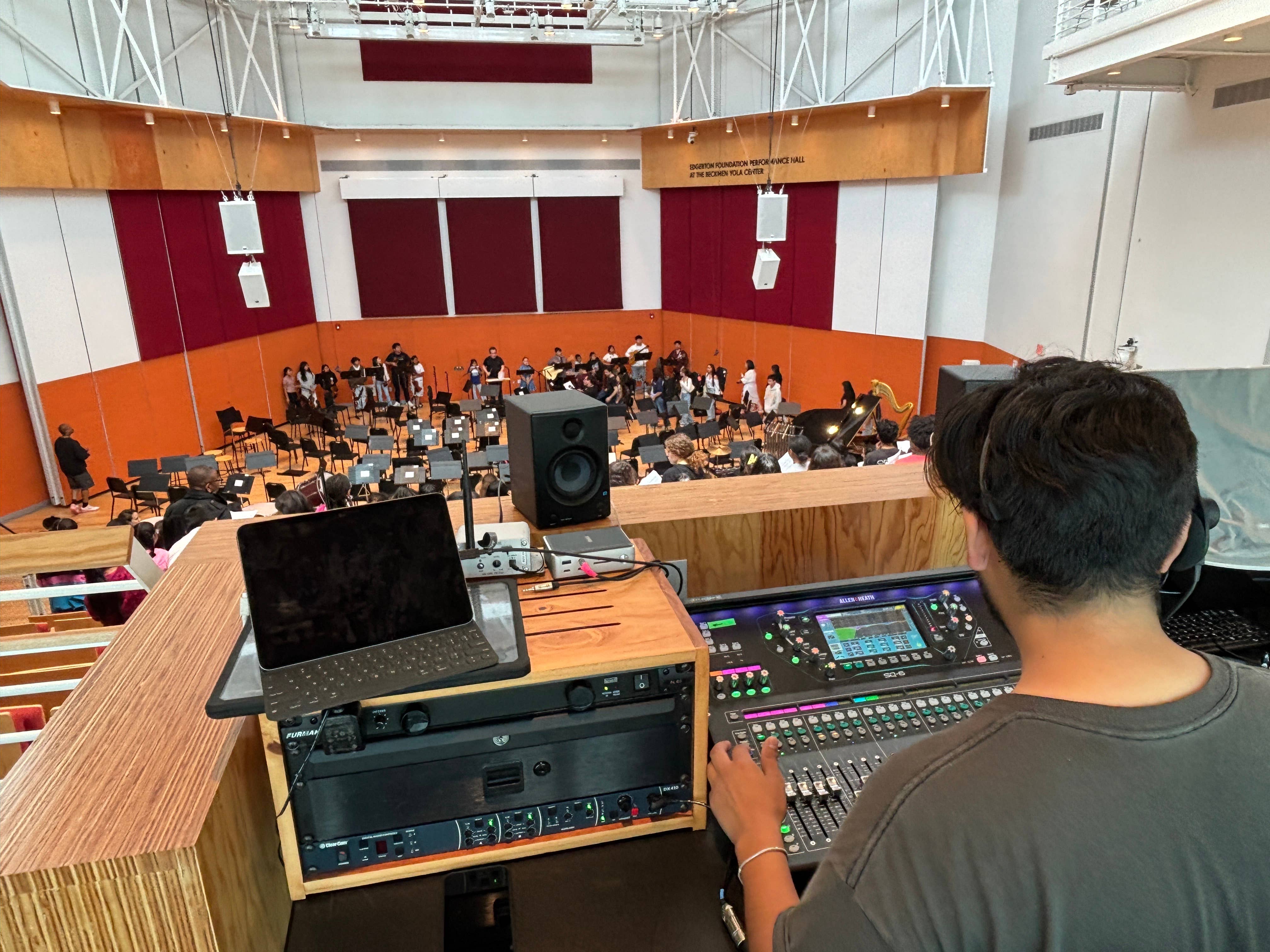 Concert hall rehearsal with a conductor and musicians on stage, while a sound engineer operates audio equipment in the foreground.