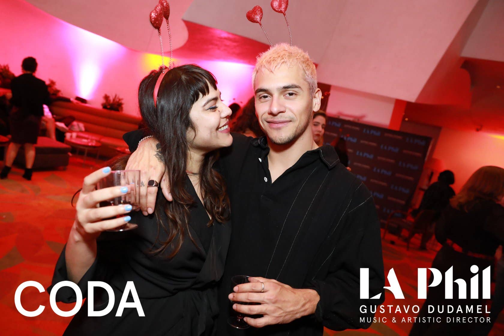 Couple at LA Phii event, woman in black holding a drink, man with pink-styled hair, red balloons overhead