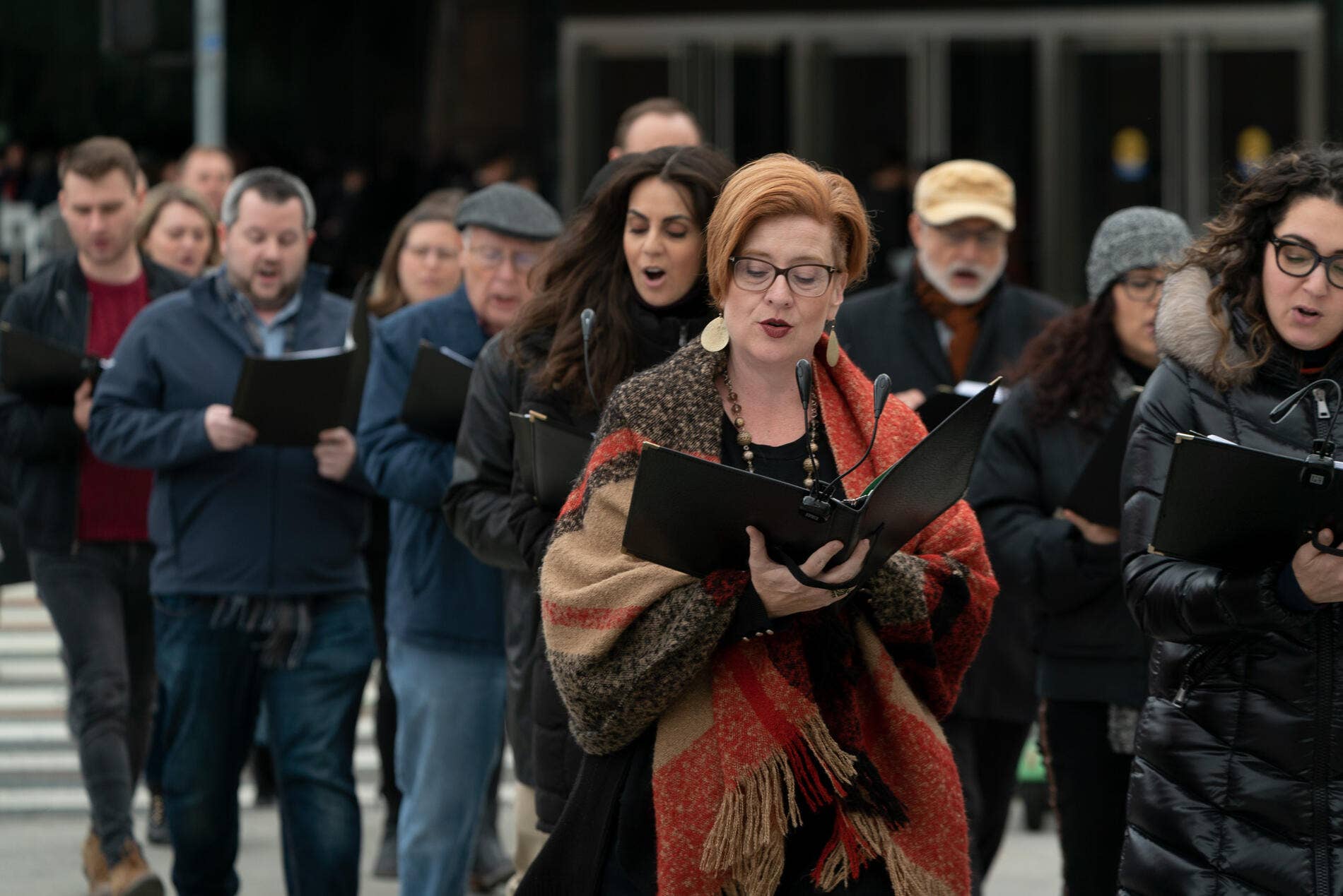 Woman in tweed coat reads from folder at outdoor podium, with crowd and security behind her.
