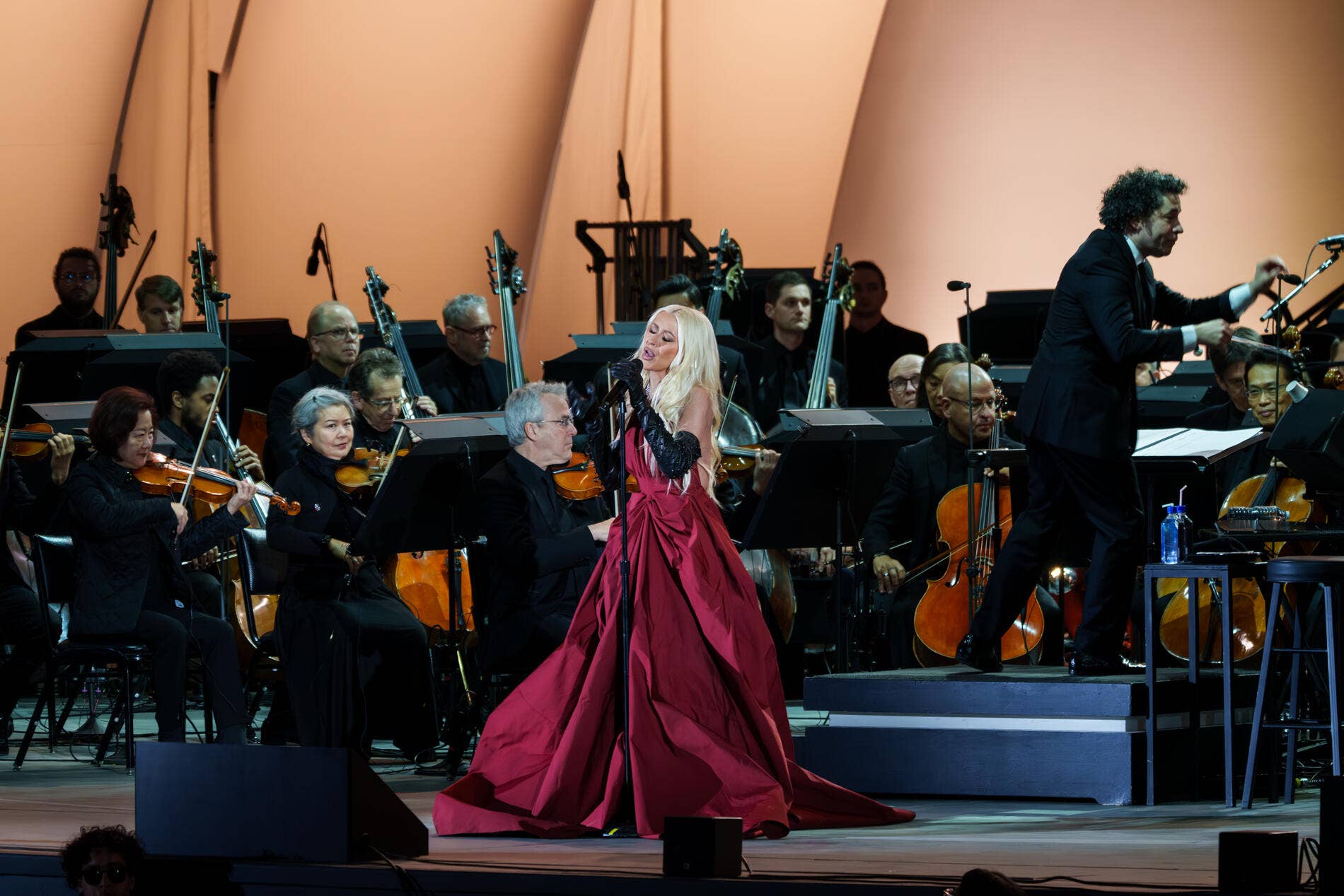 Soprano in a red gown sings at a microphone as a seated orchestra performs with strings and a guitarist.
