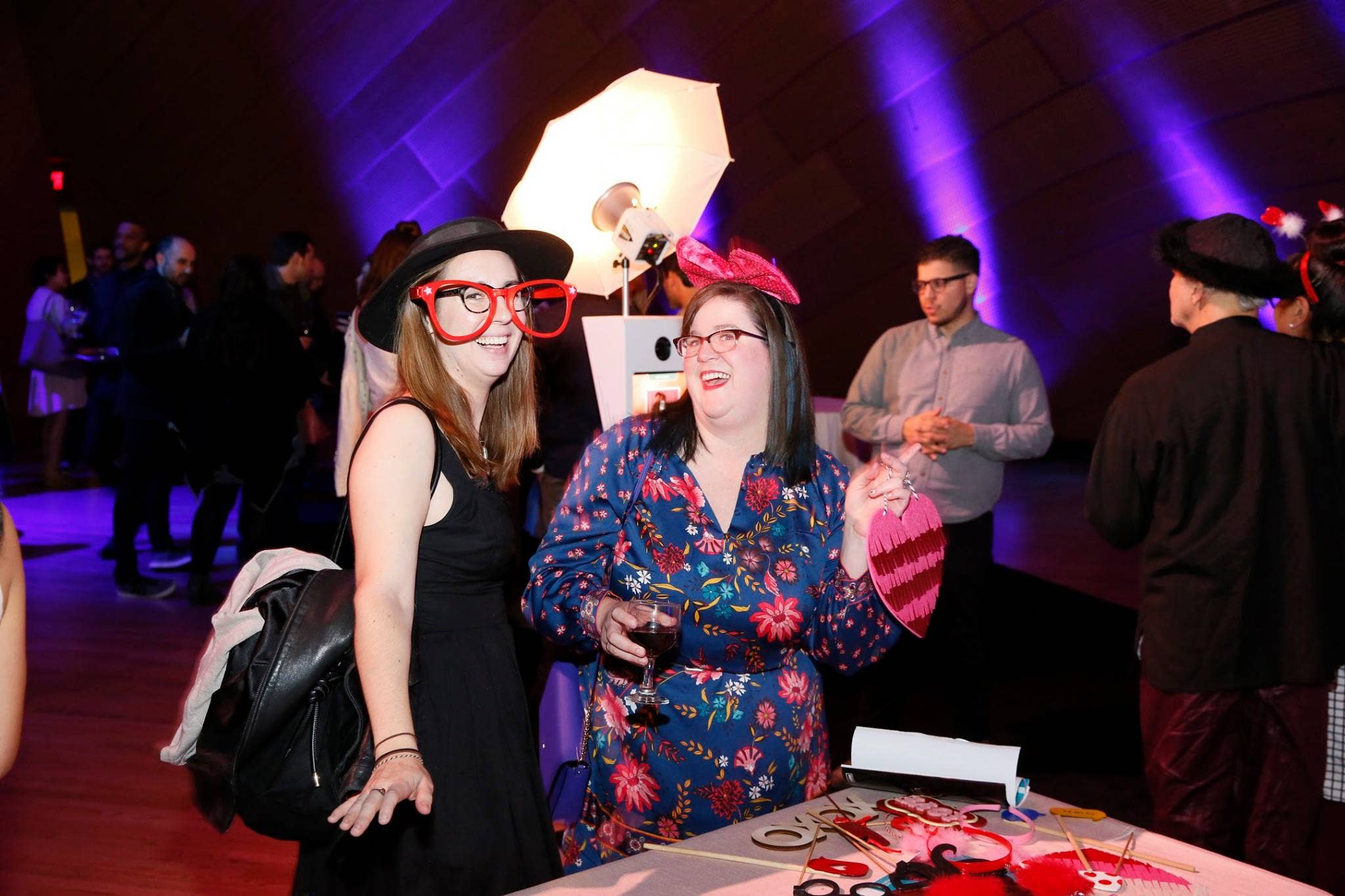 Two women in party hats and glasses smile at a table, lit by purple stage lights at an indoor event.
