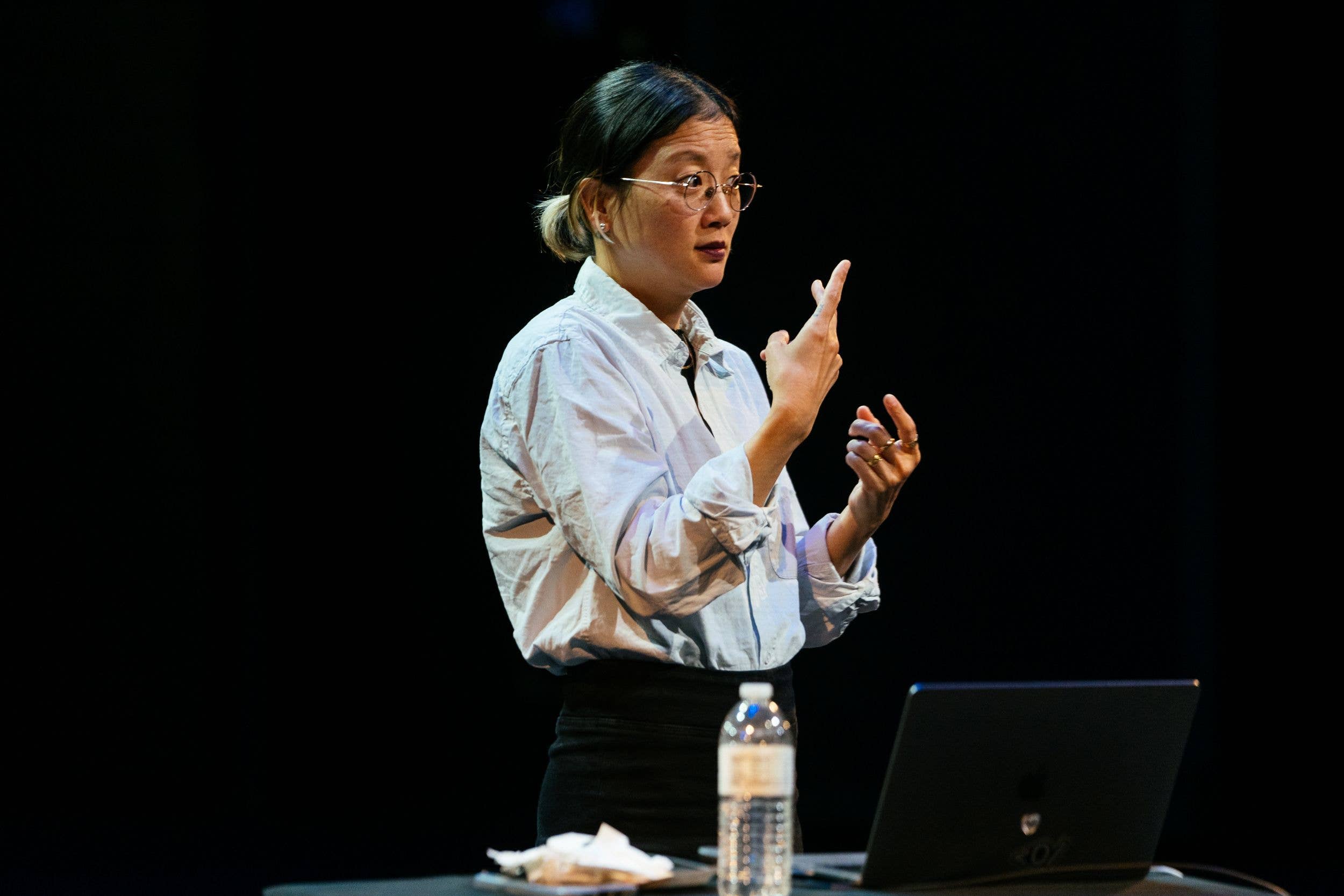 Woman in glasses speaking on stage, gesturing with raised hands beside a laptop and water bottle.
