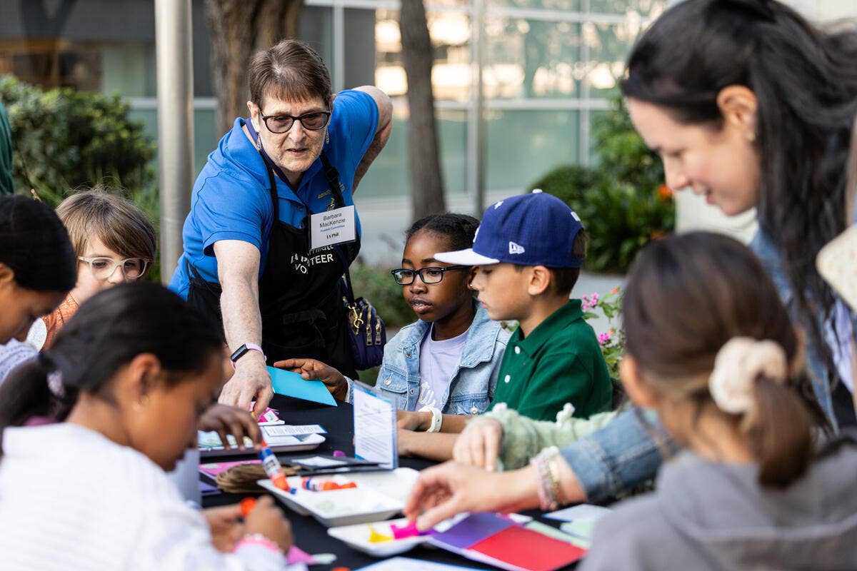 Volunteer in blue shirt teaches children at a table, pointing to materials during an outdoor activity.