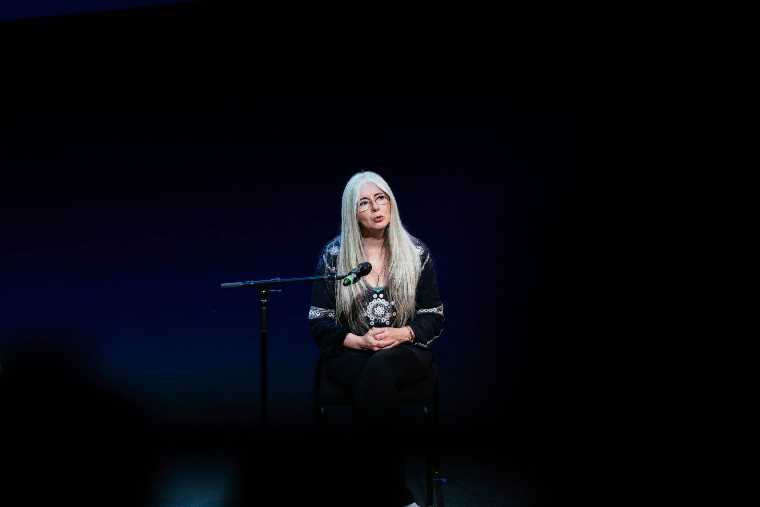 Woman with long gray hair and glasses speaks at a podium, seated on stage with a microphone against a dark blue background.