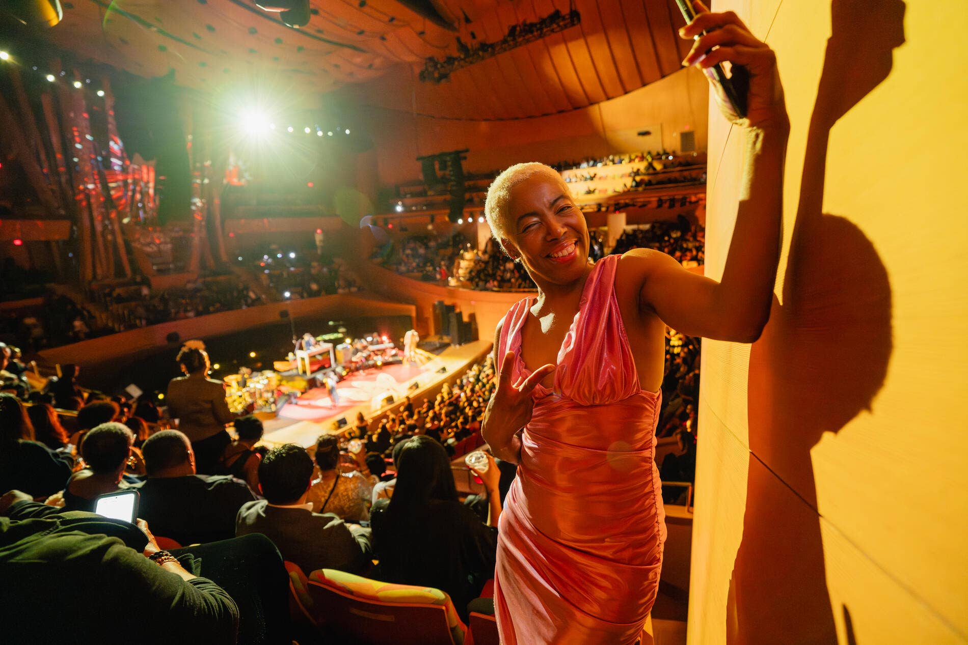 Smiling woman in an orange dress poses at a packed concert taking a selfie in warm lights