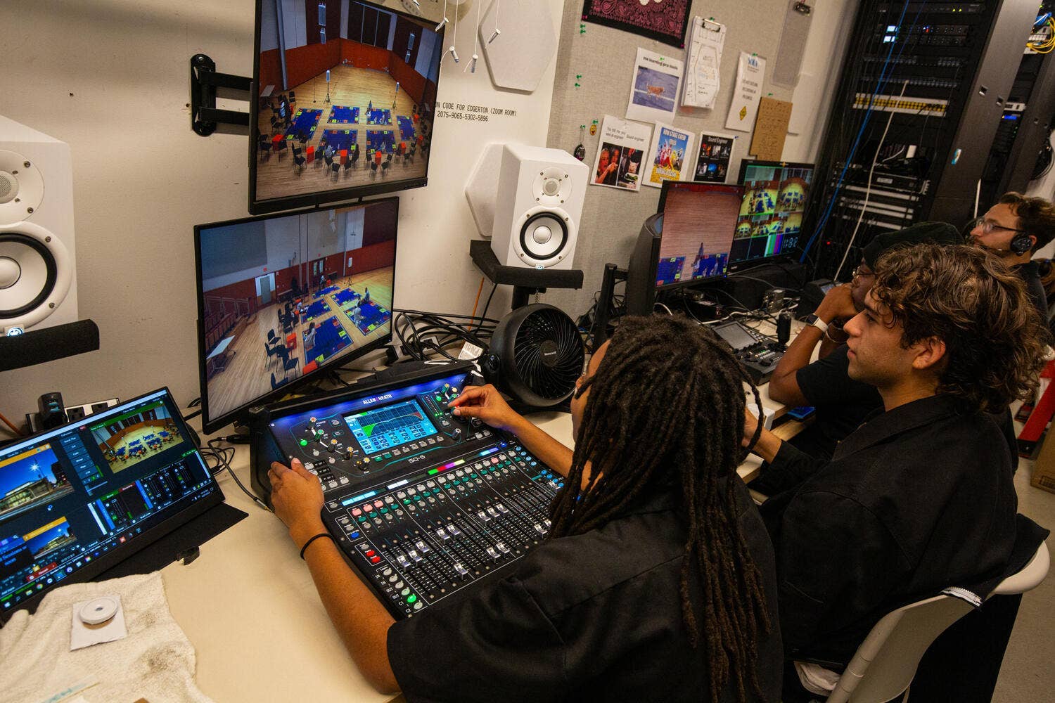 Technician operates an audio/production control console with multiple monitors and server racks in a studio control room.