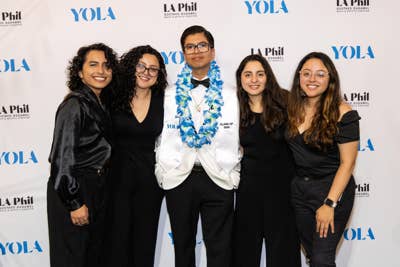 A YOLA alumni stands with family and friends in front of a step and repeat backdrop during the graduation Soirée.