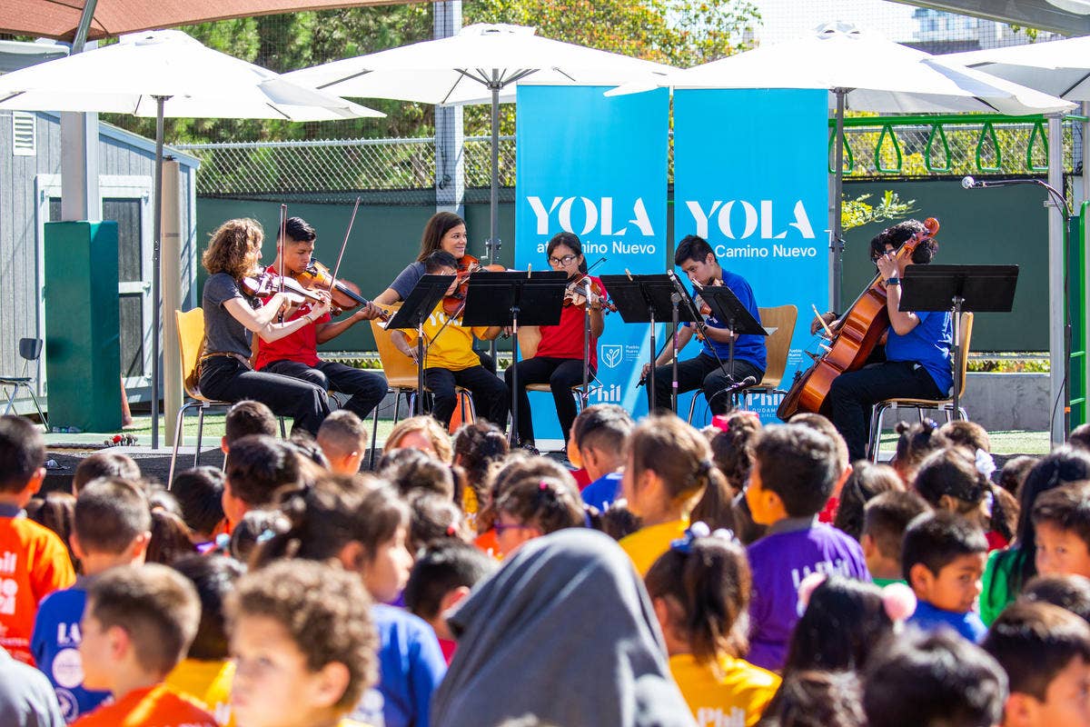 YOLA students play string instruments on a stage outdoors under umbrellas in front of an audience of other YOLA students.