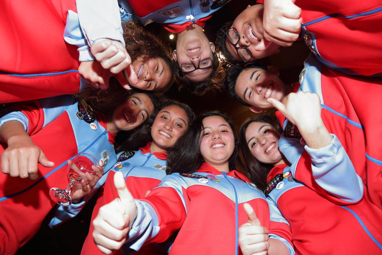 Group of people in red-and-blue team jackets posing together with thumbs-up and smiles.
