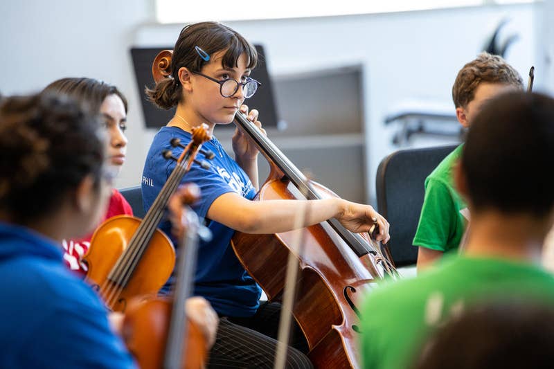 Young YOLA student plays cello in music class while classmates sit nearby in a classroom.
