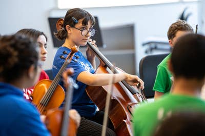Young YOLA student plays cello in music class while classmates sit nearby in a classroom.