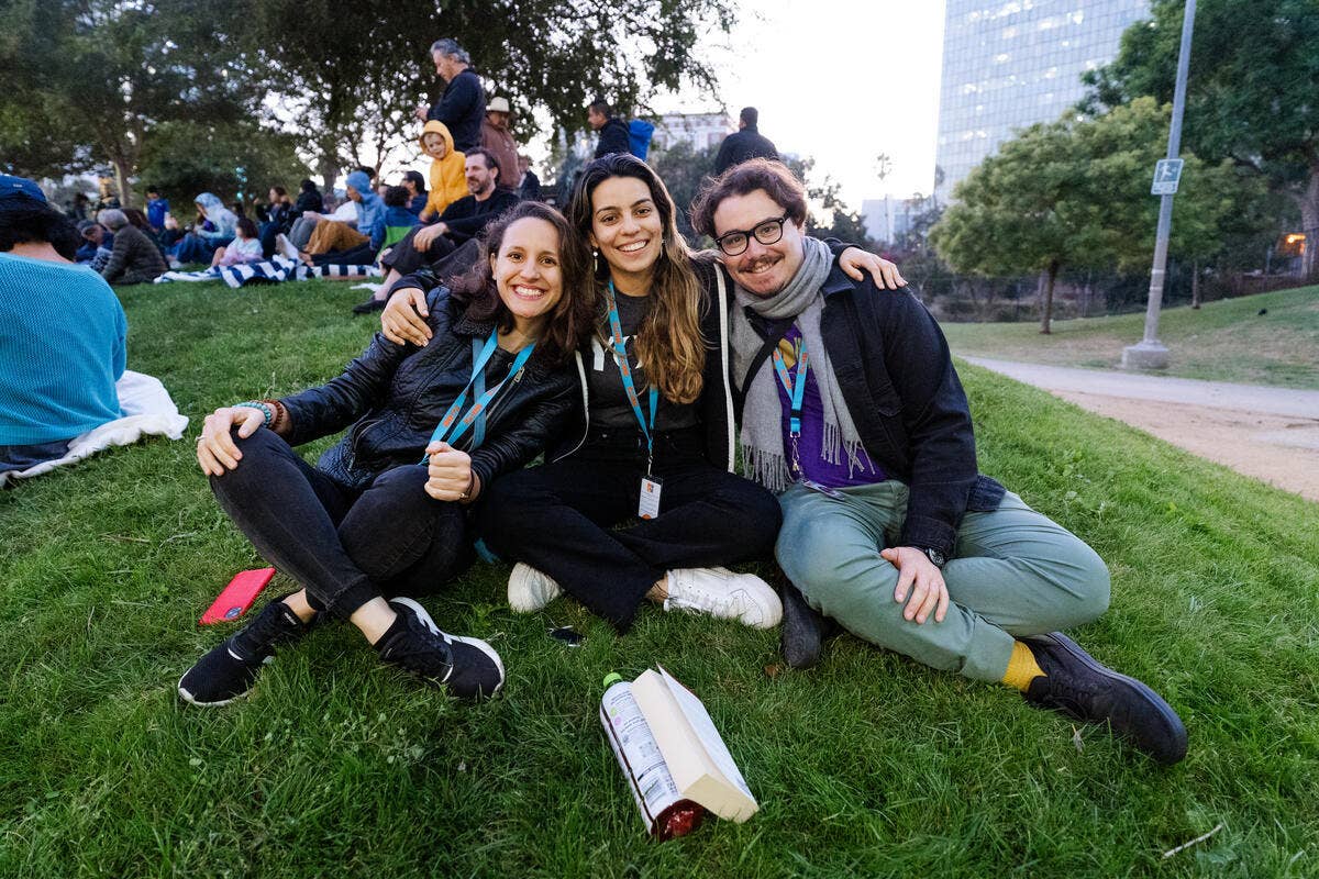 Three YOLA teaching artists embrace for a group photo while sitting on the grass outside.