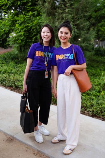 Two women smiling outdoors, wearing purple music department T-shirts against a lush green park background.