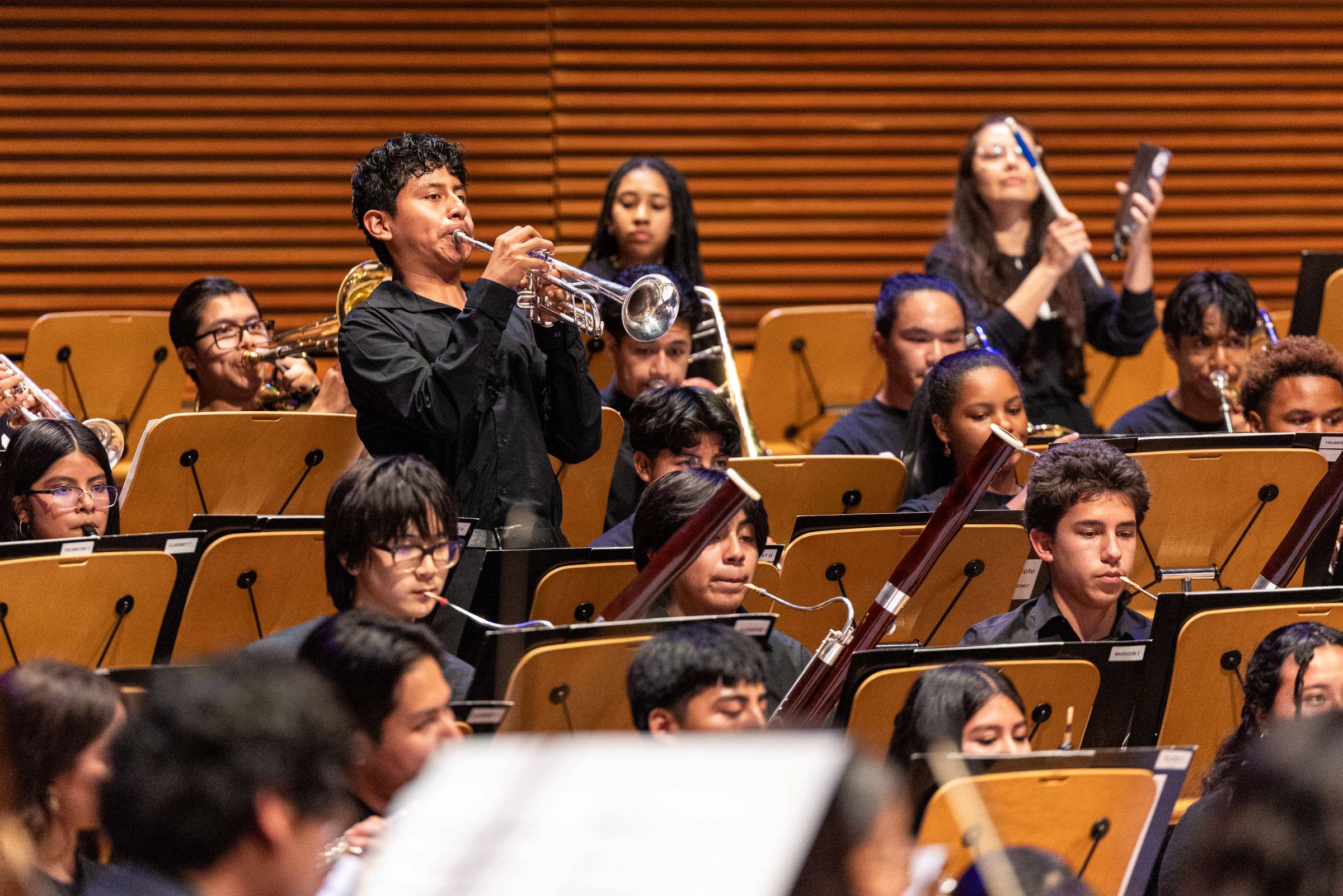 Student musicians perform in an orchestra hall, with a trumpeter and other band members holding brass and woodwinds.