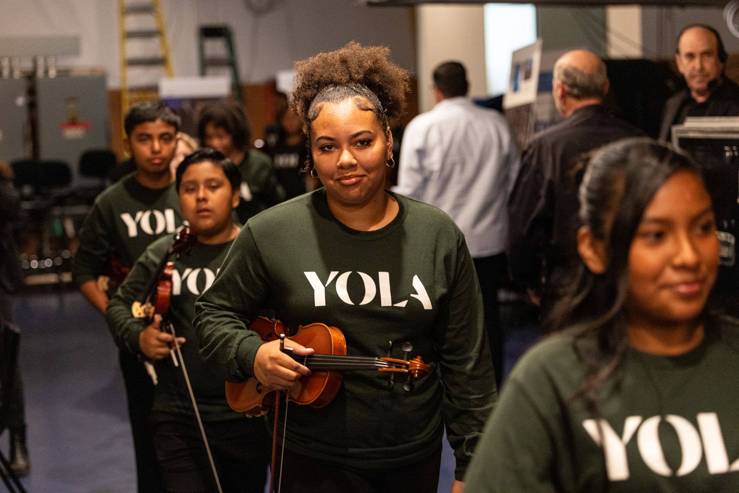 Young woman with curly hair holding a violin at a rehearsal, with other musicians in a blurred workshop background.