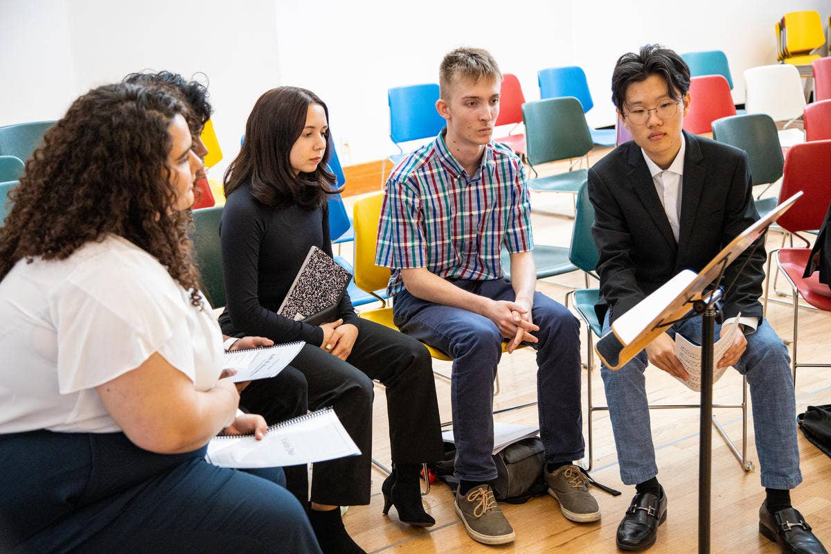 Students in a classroom listen to a speaker seated at colorful chairs, discussing notes and presentations.