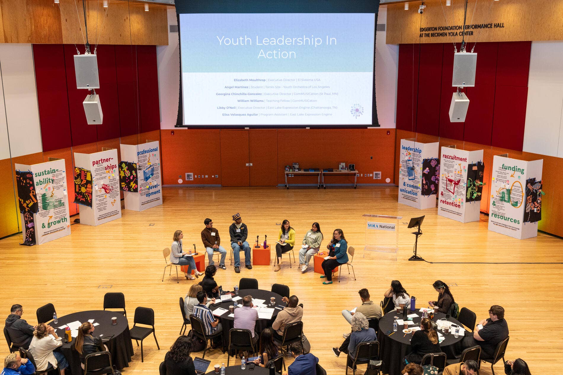 Panel discussion at “Youth Leadership in Action” conference with speakers seated on stage and audience in hall.