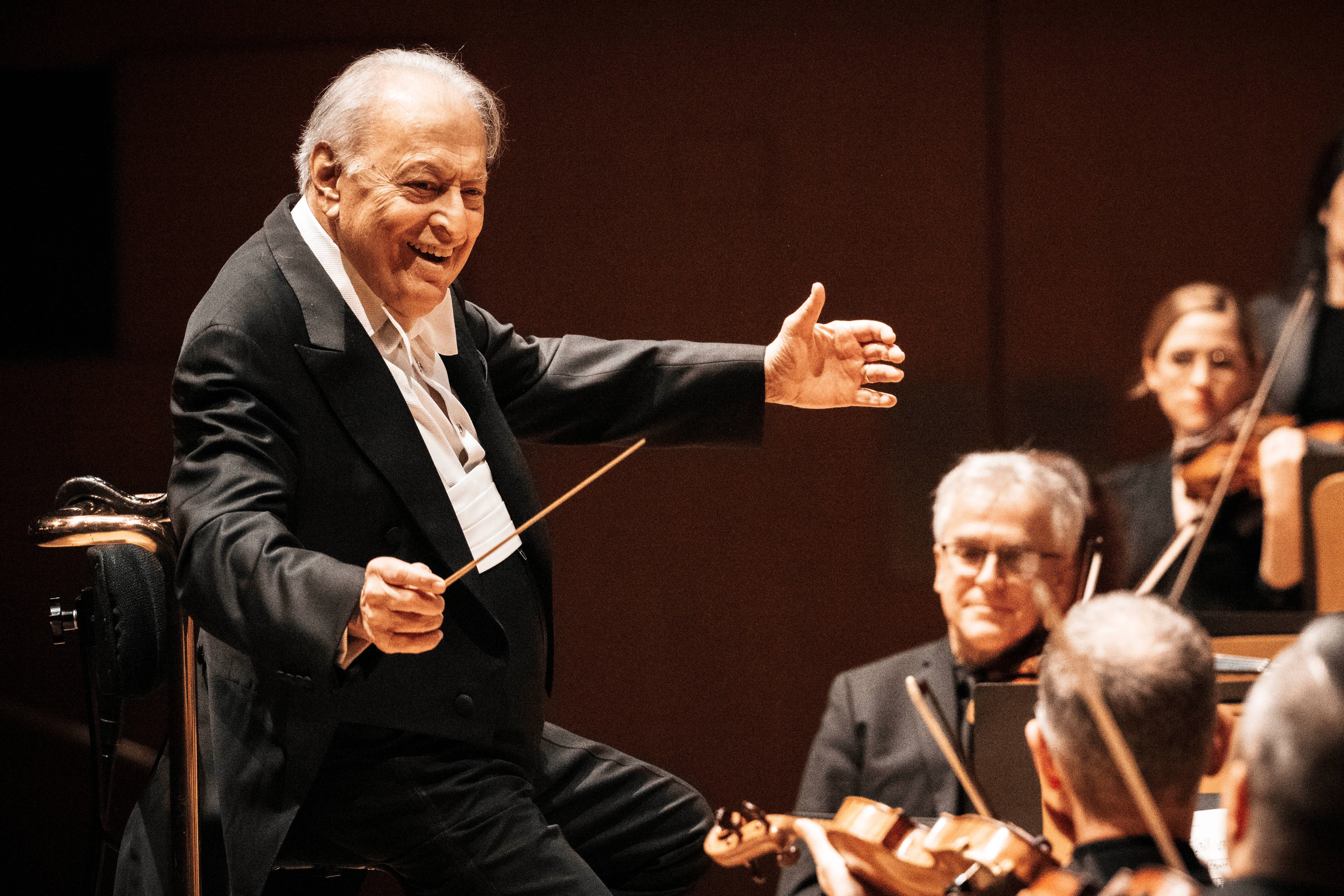 Elderly conductor in dark suit smiling and conducting an orchestra with musicians blurred in foreground and background
