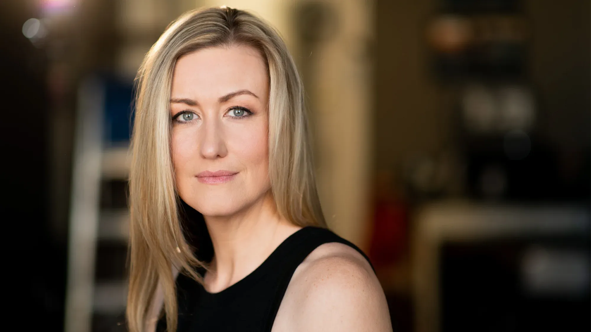Blonde woman with green eyes wearing a black top, facing the camera with a soft smile, blurred background.