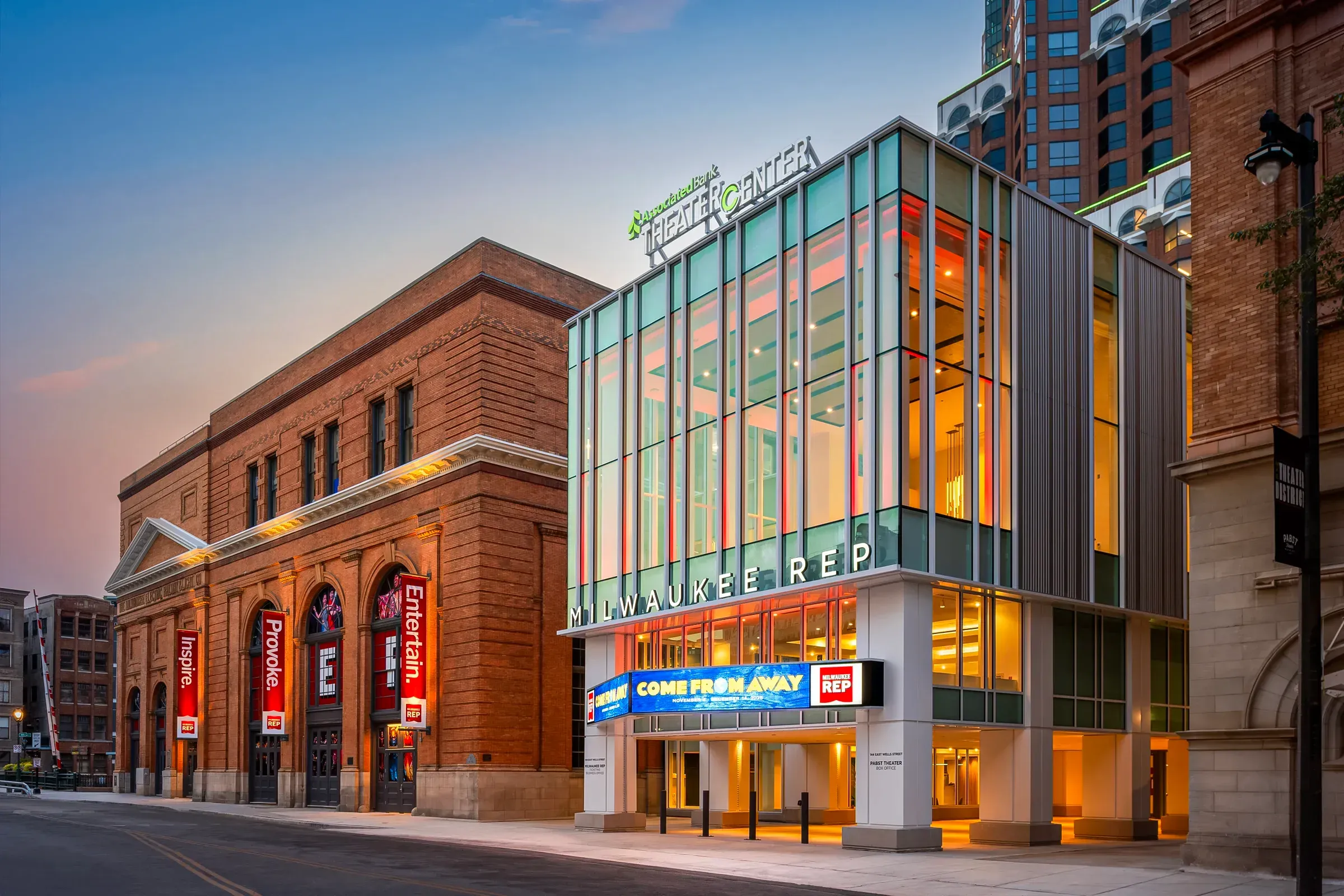 Marquee lights and glass facade outside Milwaukee Repertory Theater at dusk, with nearby brick buildings and banners