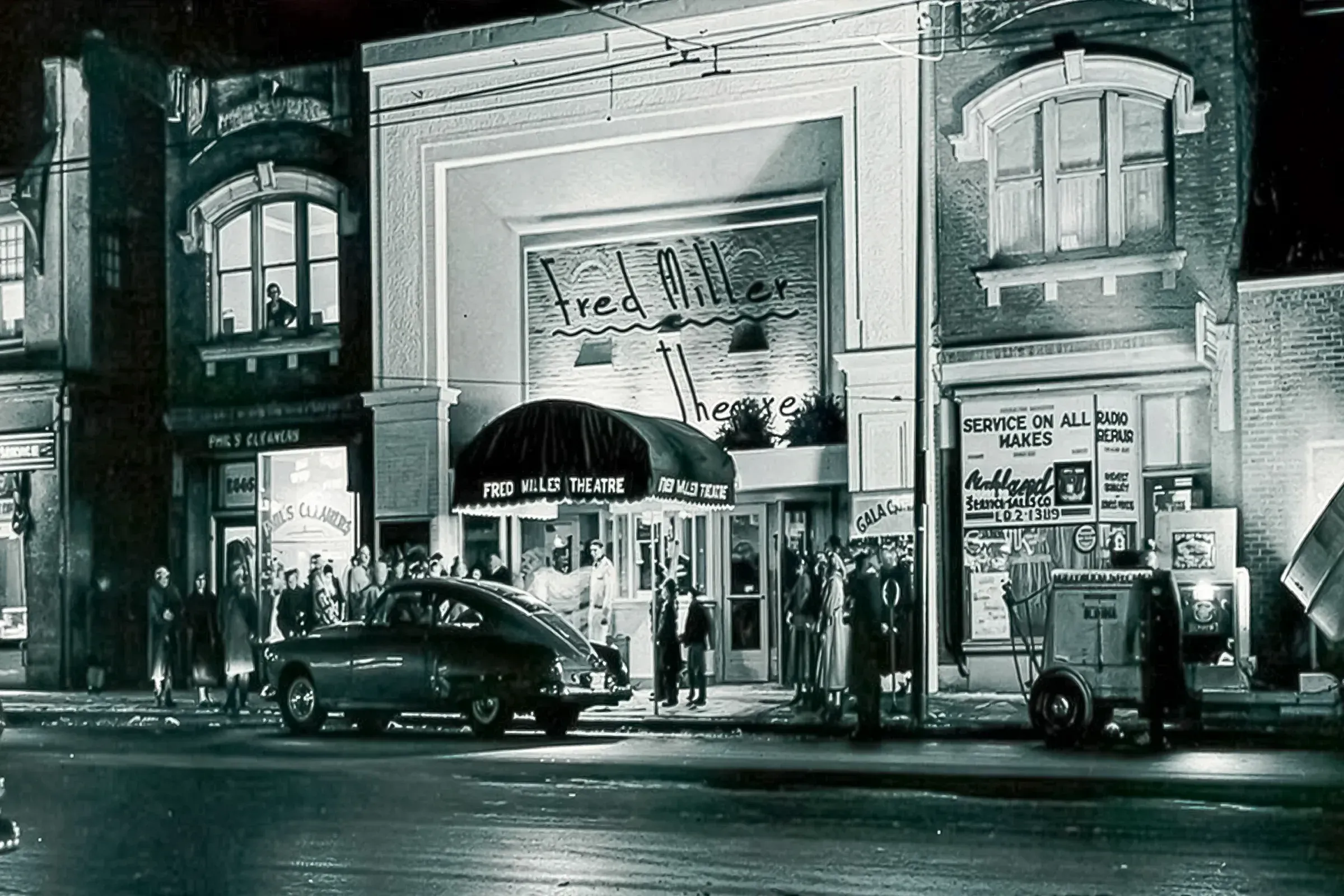 Crowd outside a theater marquee on a busy street, with vintage cars and storefront signs.