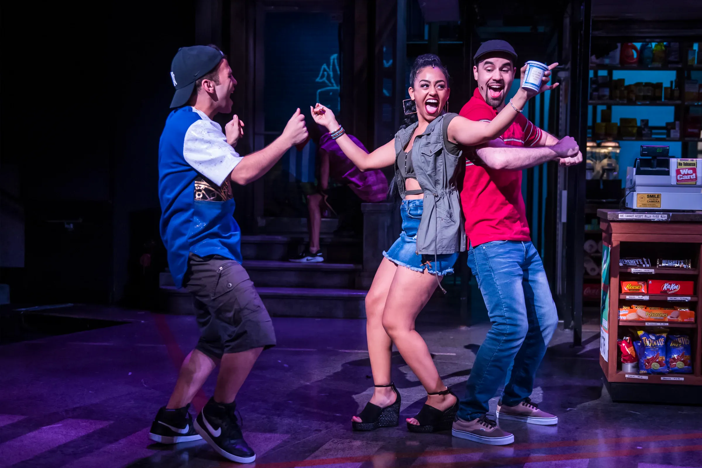 Three performers in colorful streetwear laugh and pose on stage, with one holding a can toward the viewer.