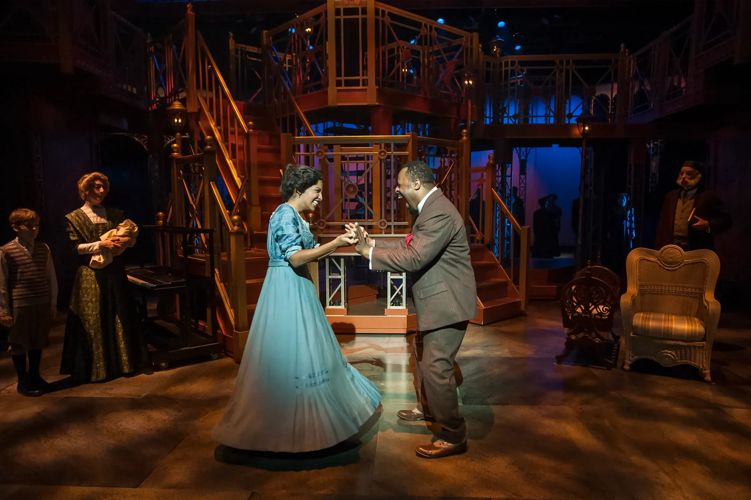 Actors in period costumes holding hands on a theater stage with ornate metal set and blue-lit backdrop at night.