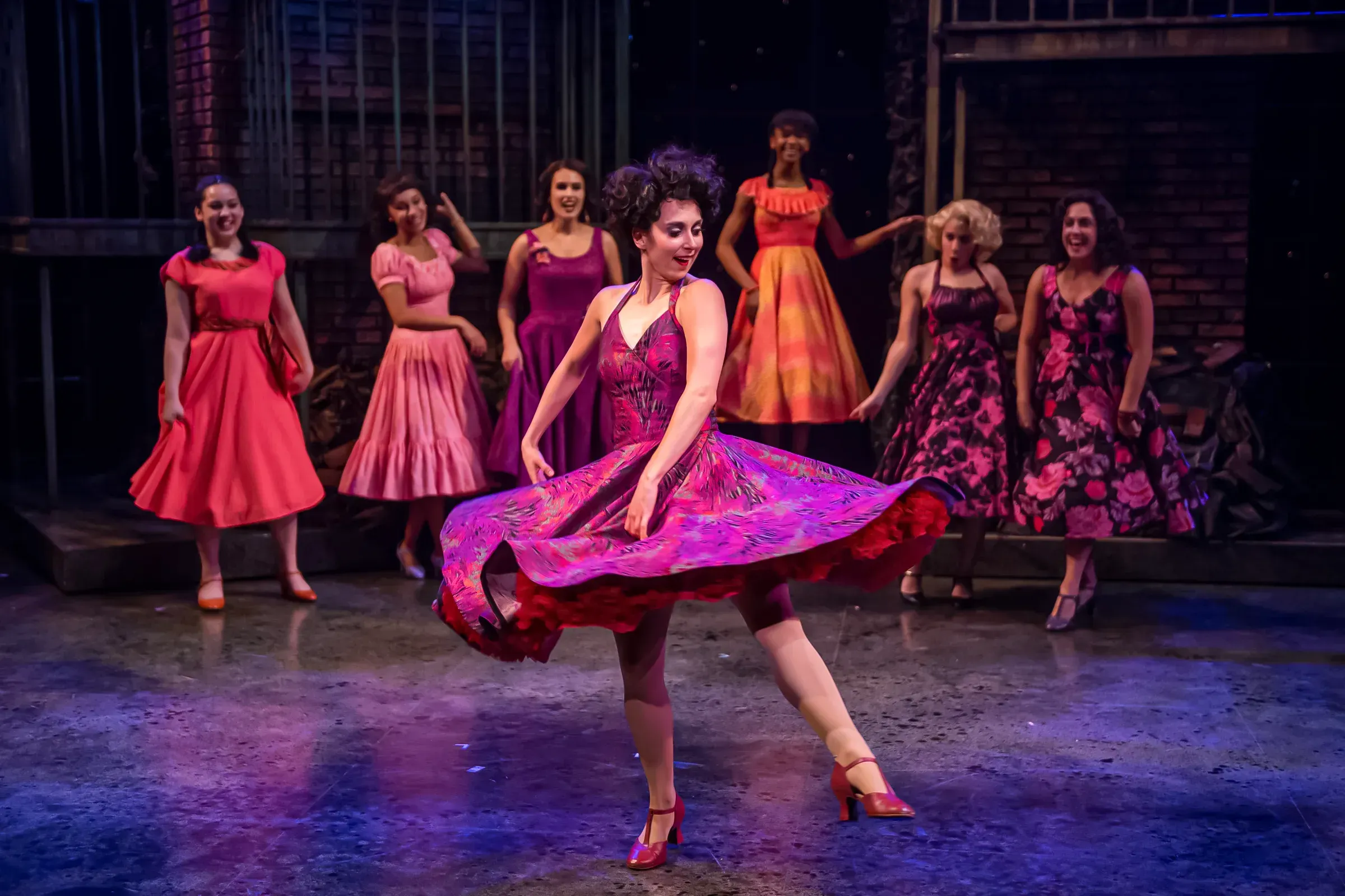 Stage dancers in colorful dresses perform a choreographed pose in a brick-walled theatre with spotlight lighting