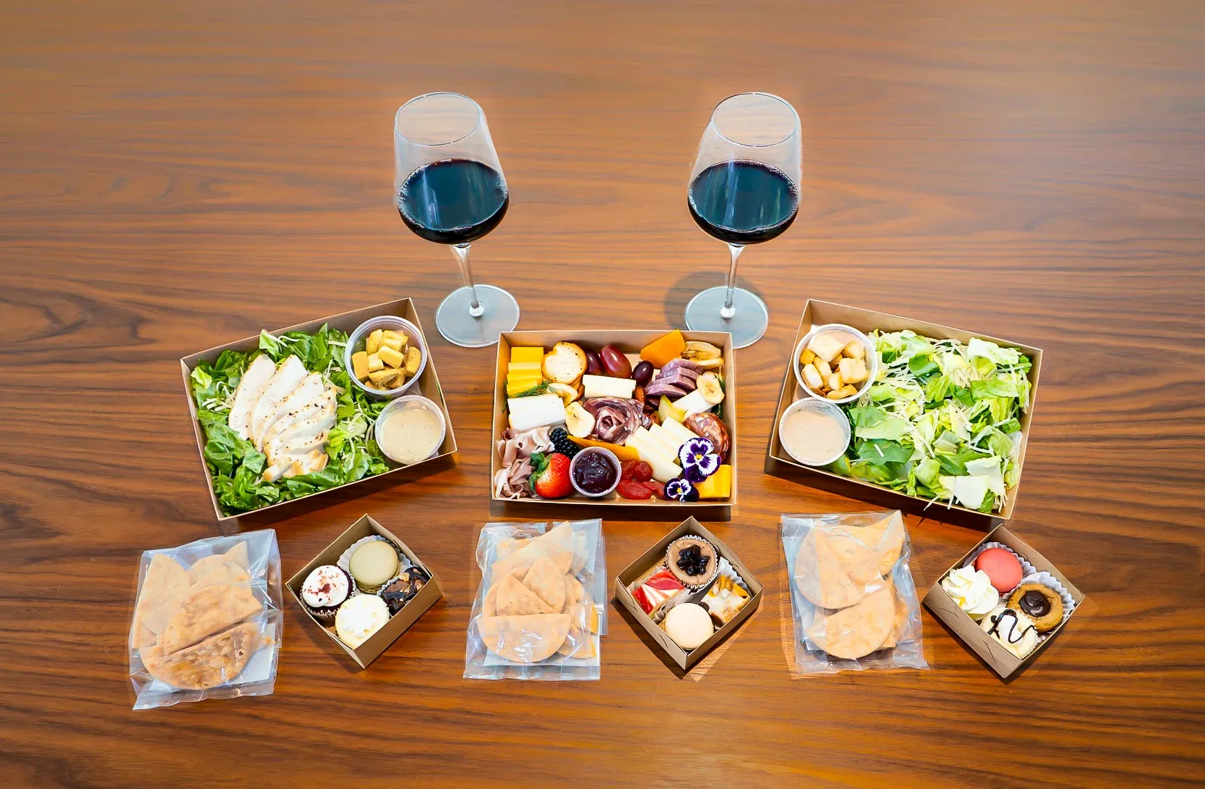 Two glasses of dark red wine beside assorted boxed salad, cheeses, charcuterie, and desserts on a wooden table.