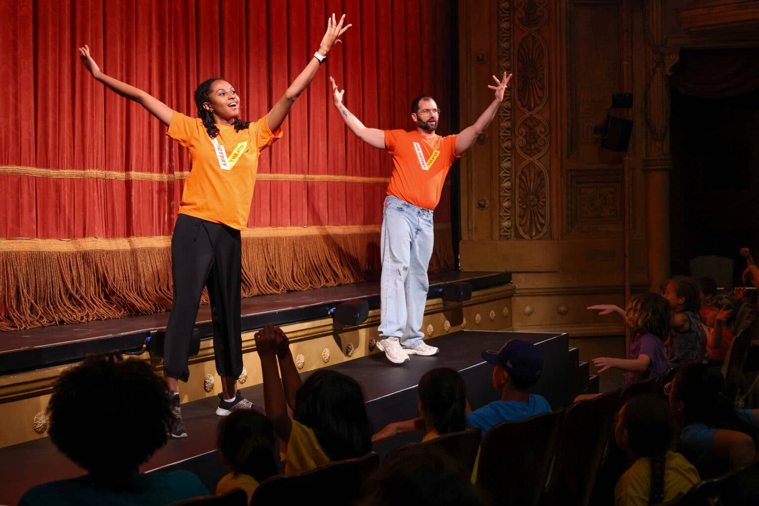 Two teaching artists in orange shirts gesture with raised arms on a theater stage before an audience.