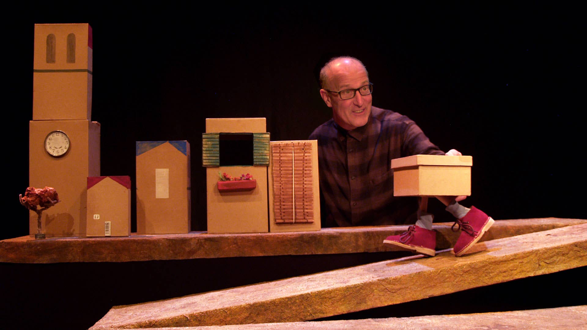 Smiling older man onstage holding a large cardboard box amid props and stacks under warm spotlight on stage.