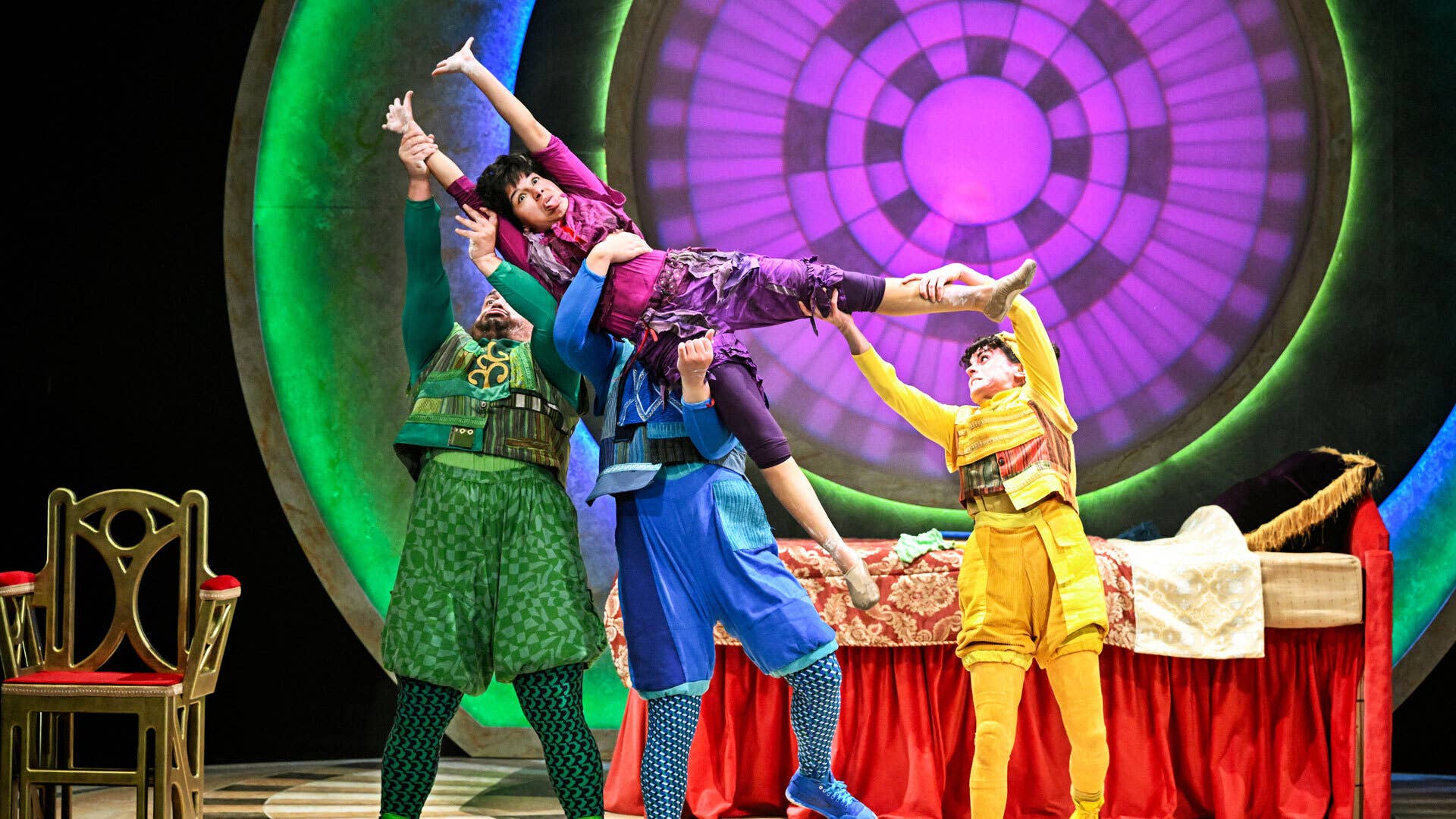 3 performers dressed in green, blue and yellow holding a performer dressed in purple up on a stage.