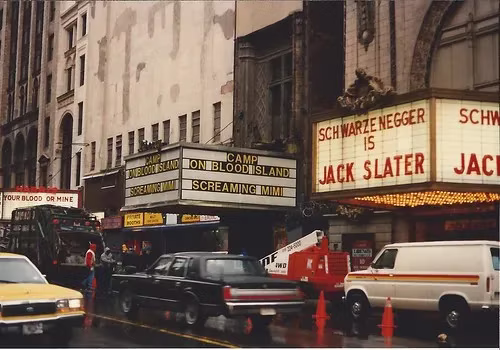Busy street with cars, billboards, and pedestrians near the Empire Theater marquee reading Schwarzenegger and Jack Slater.