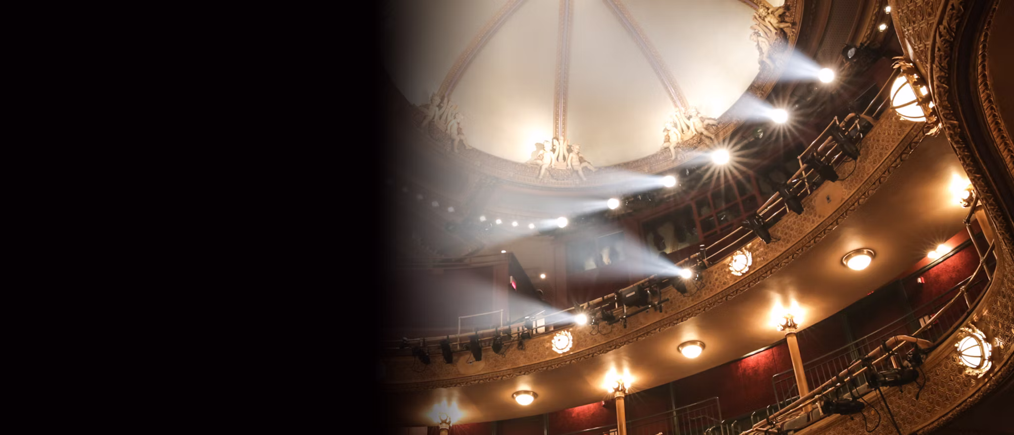 Grand theater auditorium with curved balcony, ornate gold trim, red seats, and stage spotlights.