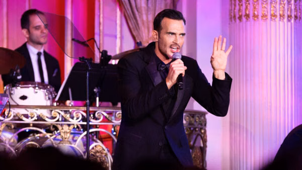 Man in a black suit sings into a microphone while gesturing at an event with stage lights and brass decor.