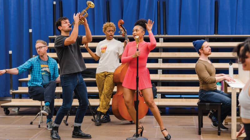 Performers rehearsing on stage with trumpet, saxophone, and microphone under blue curtains.