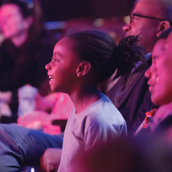 A young girl sitting in the audience smiling towards the stage. 