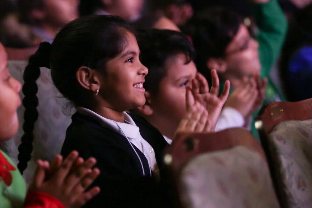 A young girl in a dark blazer applauds with a cheering crowd in a brightly lit auditorium.