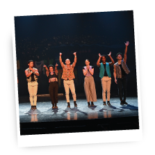 Group of dancers performing on stage under colorful lights, posing with arms raised