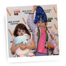 Two smiling children pose in front of a colorful backdrop with “New Victory” signs.