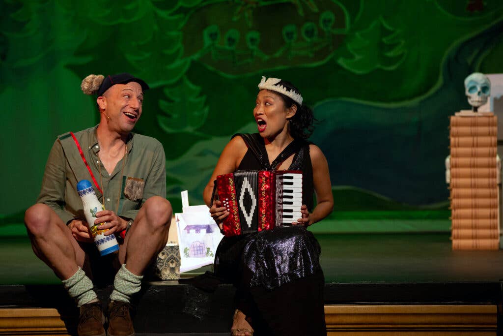 Anton Dudley and Faye Chiao sit on the edge of the stage singing while Faye plays accordion.