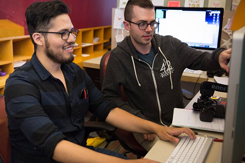 Two men sit at desks with computers, one smiling while pointing at the monitor in an office.